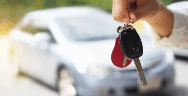 Close-up of a person’s hand holding car keys with a blurred silver car in the background, symbolizing buying, selling, or renting a vehicle