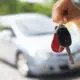 Close-up of a person’s hand holding car keys with a blurred silver car in the background, symbolizing buying, selling, or renting a vehicle