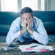 Stressed man covering his face with both hands while sitting in front of a calculator and scattered bills on a coffee table.