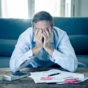 Stressed man covering his face with both hands while sitting in front of a calculator and scattered bills on a coffee table.