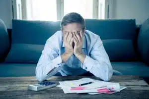 Stressed man covering his face with both hands while sitting in front of a calculator and scattered bills on a coffee table.