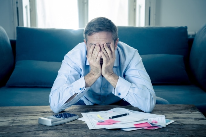 Stressed man covering his face with both hands while sitting in front of a calculator and scattered bills on a coffee table.