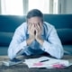 Stressed man covering his face with both hands while sitting in front of a calculator and scattered bills on a coffee table.
