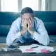 Stressed man covering his face with both hands while sitting in front of a calculator and scattered bills on a coffee table.