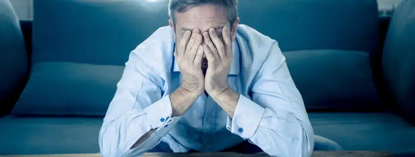 Stressed man covering his face with both hands while sitting in front of a calculator and scattered bills on a coffee table.