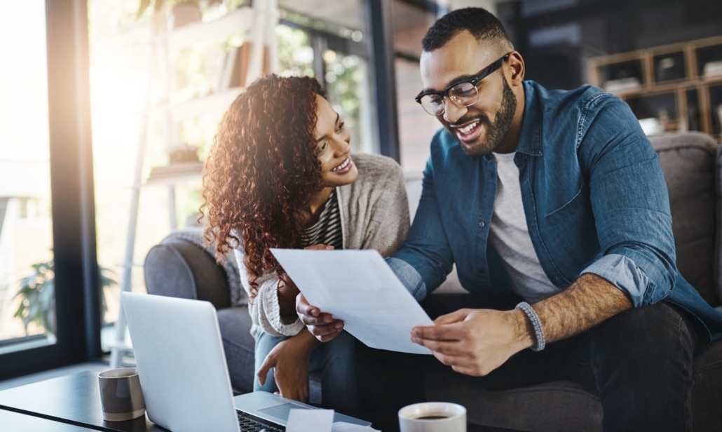 Smiling couple sitting on a couch reviewing a financial document together with a laptop and coffee on the table in front of them. 