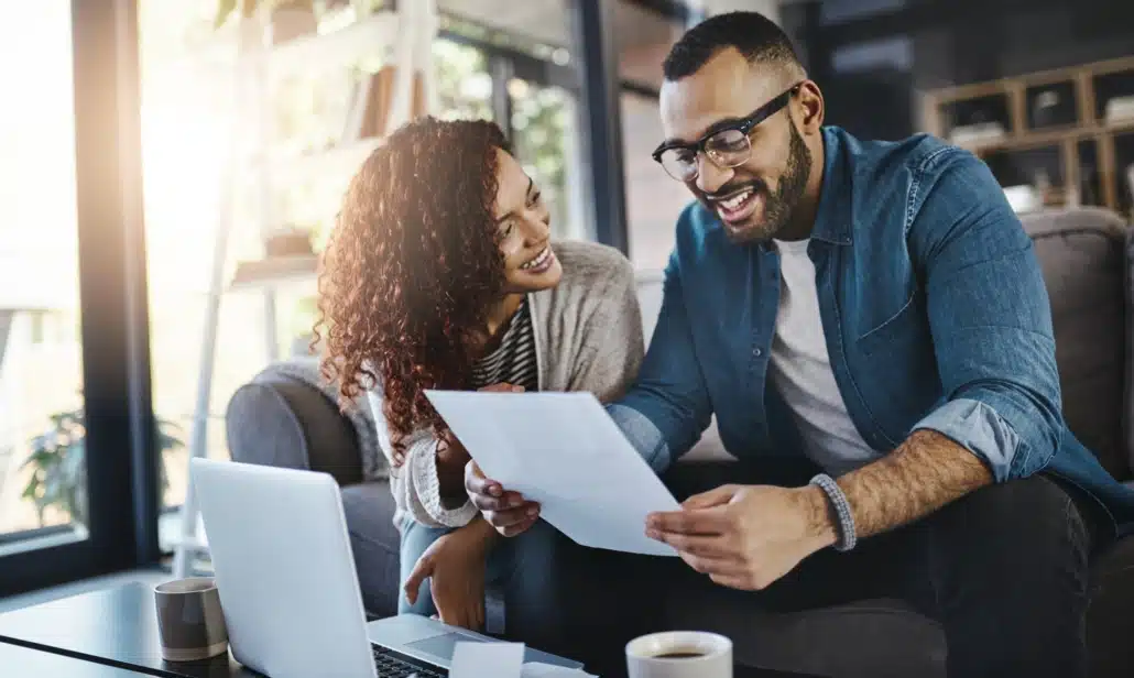 Smiling couple sitting on a couch reviewing a financial document together with a laptop and coffee on the table in front of them.