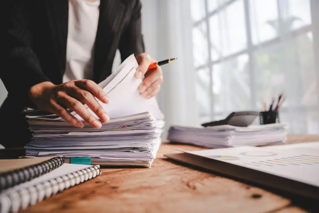 Professional sorting through a large stack of documents at a desk with a pen in hand, alongside a calculator and financial charts.