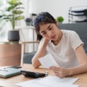 Young woman sitting at her kitchen table reviewing a tax bill with concern, surrounded by paperwork and a calculator.