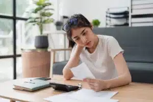 Young woman sitting at her kitchen table reviewing a tax bill with concern, surrounded by paperwork and a calculator.