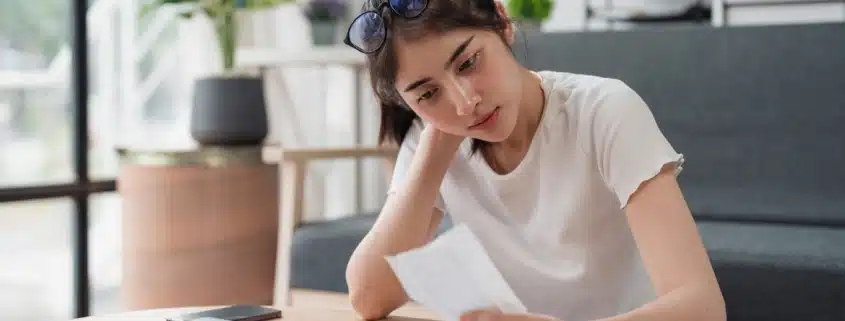 Young woman sitting at her kitchen table reviewing a tax bill with concern, surrounded by paperwork and a calculator.