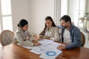 Three professionals reviewing financial analysis documents and charts around a wooden table, including forms, bar graphs, and blue pie charts