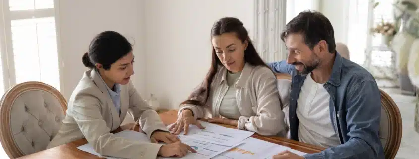 Three professionals reviewing financial analysis documents and charts around a wooden table, including forms, bar graphs, and blue pie charts