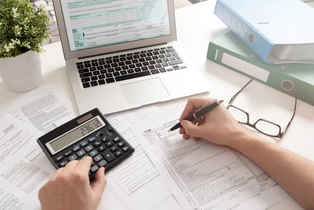 Person working on Maryland service tax documents, using a calculator to perform calculations, with tax forms and documents spread across a desk, alongside a laptop displaying a tax form on screen