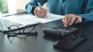 Person using a calculator while writing notes at a desk with a laptop and glasses nearby, focused on financial calculations.
