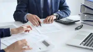 Two professionals reviewing and pointing at financial or legal documents on a desk with a calculator, laptop, and paperwork.